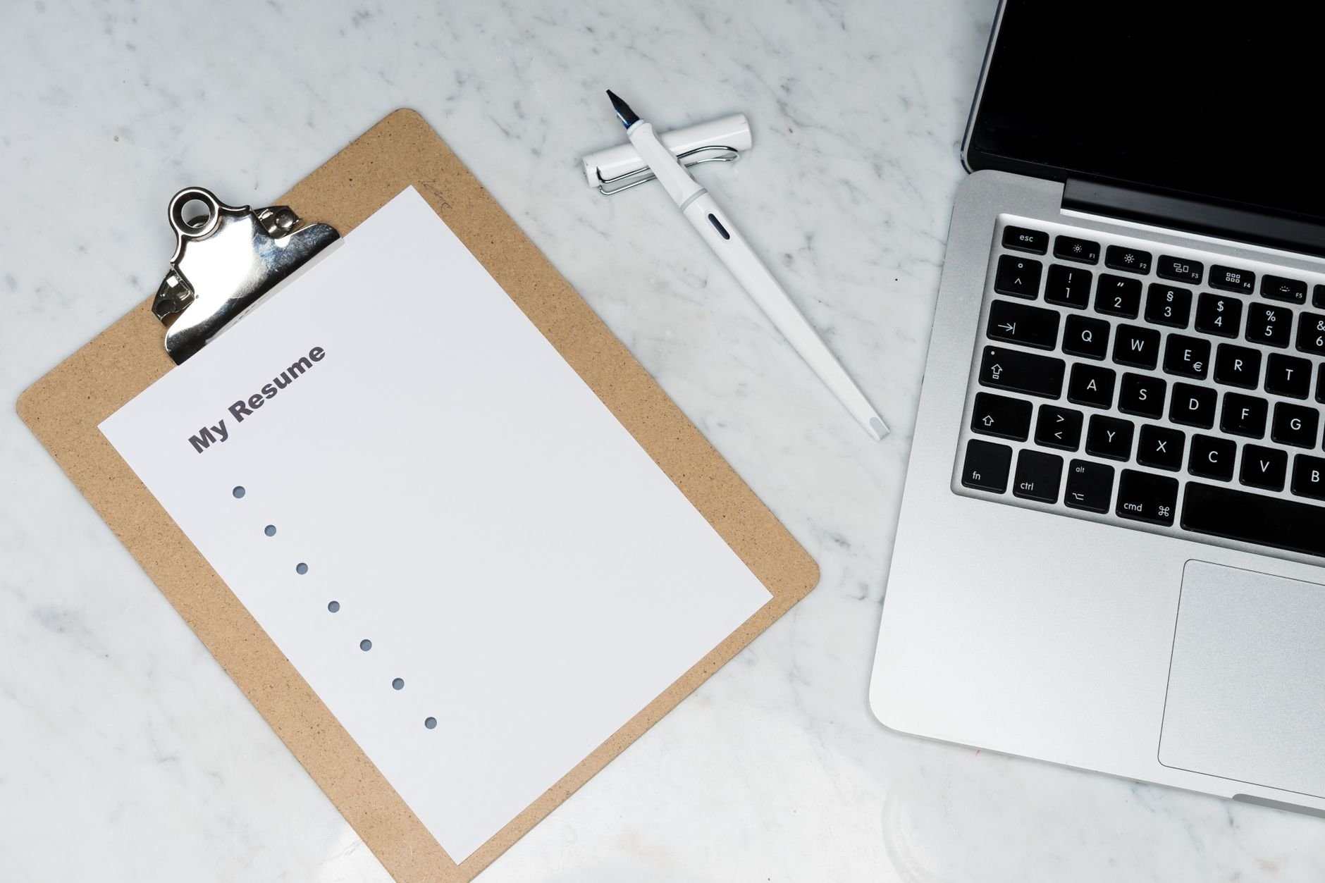 A clipboard with a resume beside a pen and laptop on a marble desk